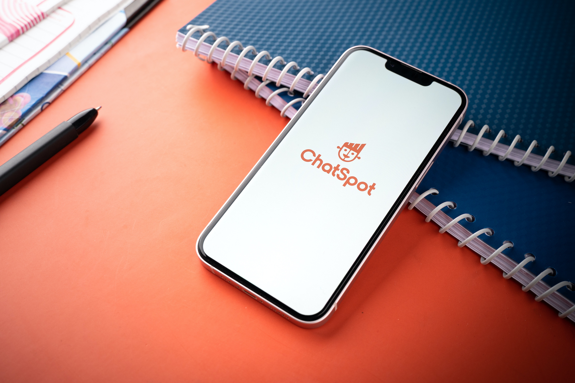 ChatSpot logo featured on a mobile device, surrounded by black notebooks and pens on an orange background